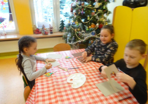 Troje dzieci siedzi przy stole, obrysowują swoje dłonie na kartce i wycinają szablony.